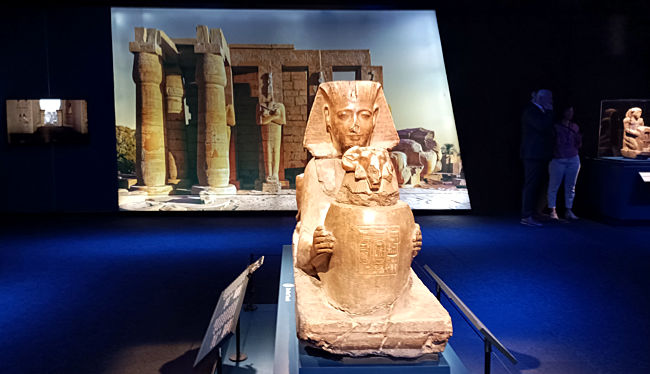 Odysseum, Ausstellung, Statue von Ramses II. als Sphinx, die ein Gefäß  mit Widderkopf darbringt, Neues Reich, Ägyptisches Museum. In dieser Sphinx, halb Löwe, halb Mensch, sind die absolute Macht und die religiöse Hingabe von Ramses verkörpert. Damals wie heute sind Löwen als Symbol physischer Kraft eng mit dem Königtum verbunden. Hier bietet Ramses dem König der Götter, Amun-Re, ein Gefäß mit heiligem Wasser an. Diese Sphinx stand im großen Tempel des Amun-Re in Karnak. Hintergrundbild: Ramesseum.