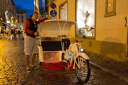 Lettland - junger Mann an seiner weißen Luxus-Fahrradrikscha in der Altstadt von Riga bei Nacht